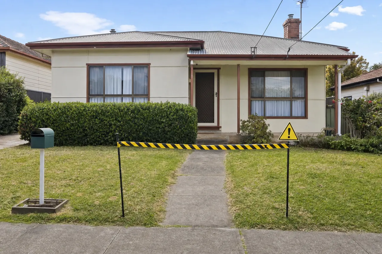 A single-storey cream house with a hipped corrugated roof behind a hedge and lawn, yellow-and-black tape strung between two stakes across the concrete front path, with a yellow triangular warning sign beside it — asbestos removal, Newcastle. Illustrative example.